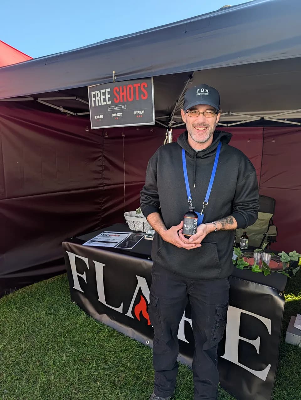 A smiling man in a black cap and hoodie holds a bottle of Flare in both hands at the stall.