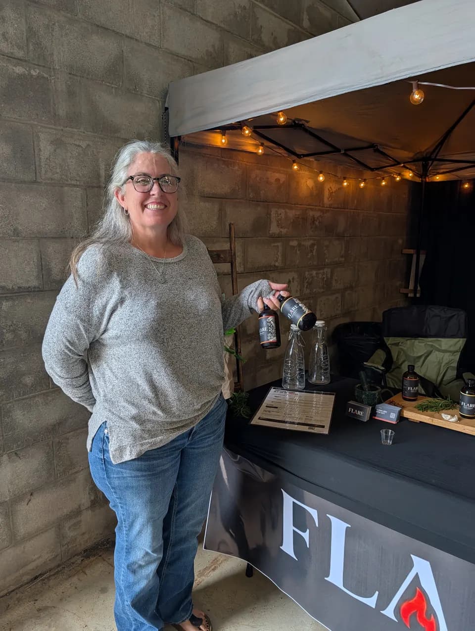A woman with long grey hair and glasses smiles beside the Flare stall, holding two bottles.