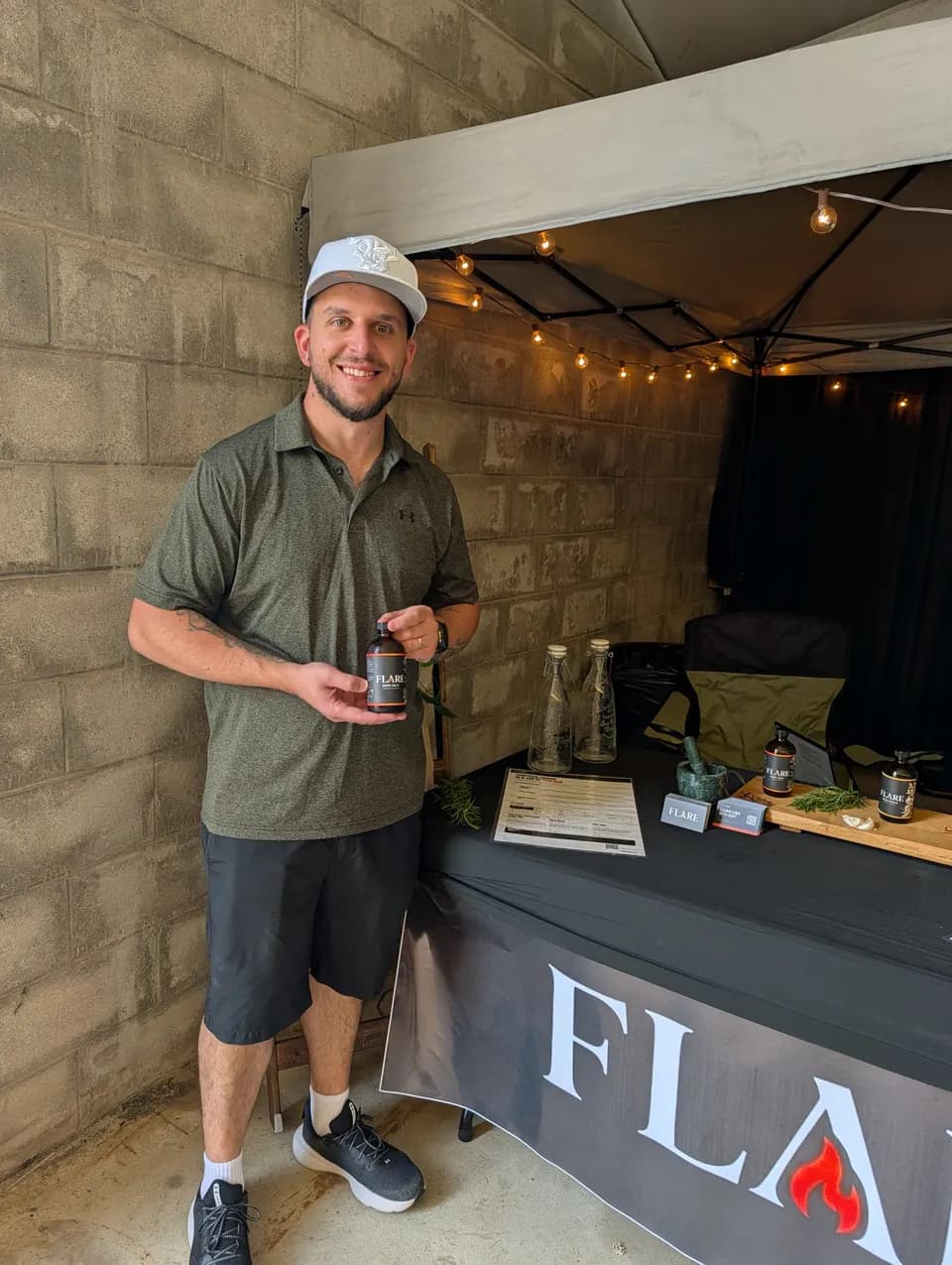 A man in a white cap and green polo holds a bottle of Flare in front of the stall.