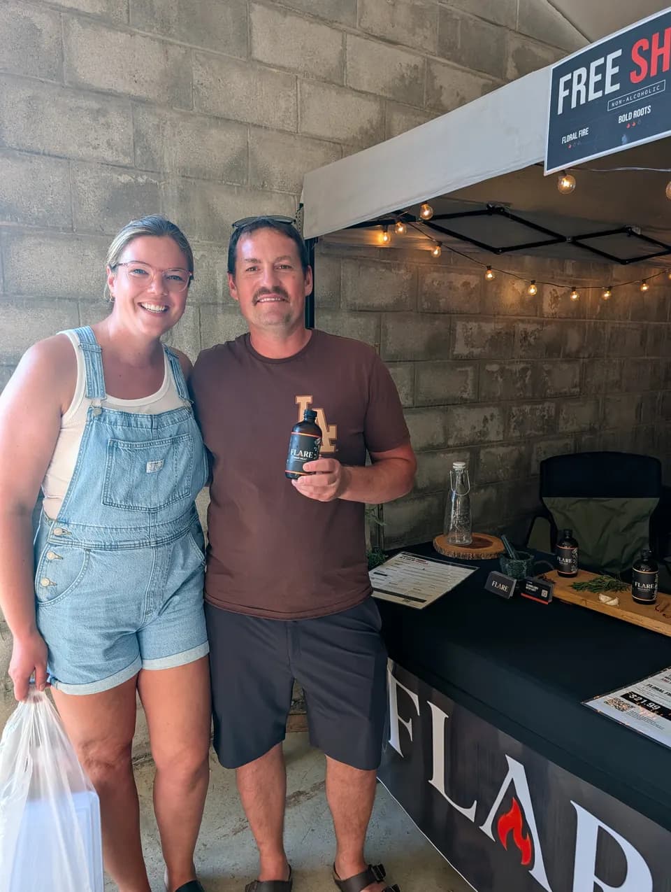 A couple stand together at the Flare stall, him holding a bottle, her carrying a bag.